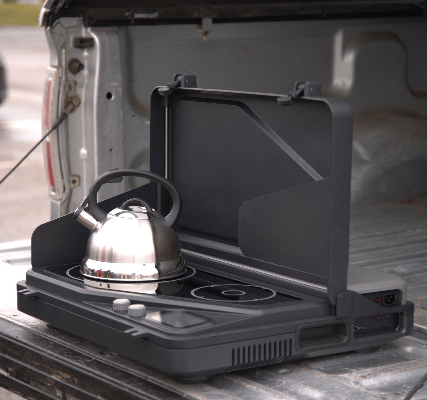 Our battery powered, portable induction cooking stove and generator, on a tailgate, for tailgating, camping, overlanding, and off-grid survival. a removable battery and dual burners for campers, outdoor workers, victims of natural disasters — and anyone, anywhere, cooking or surviving off-grid. Pictured here with the lid open with a kettle and also a coffee maker on with the dual burner system is shown in the portable Overlander model. Wind guards are currently pictured also.