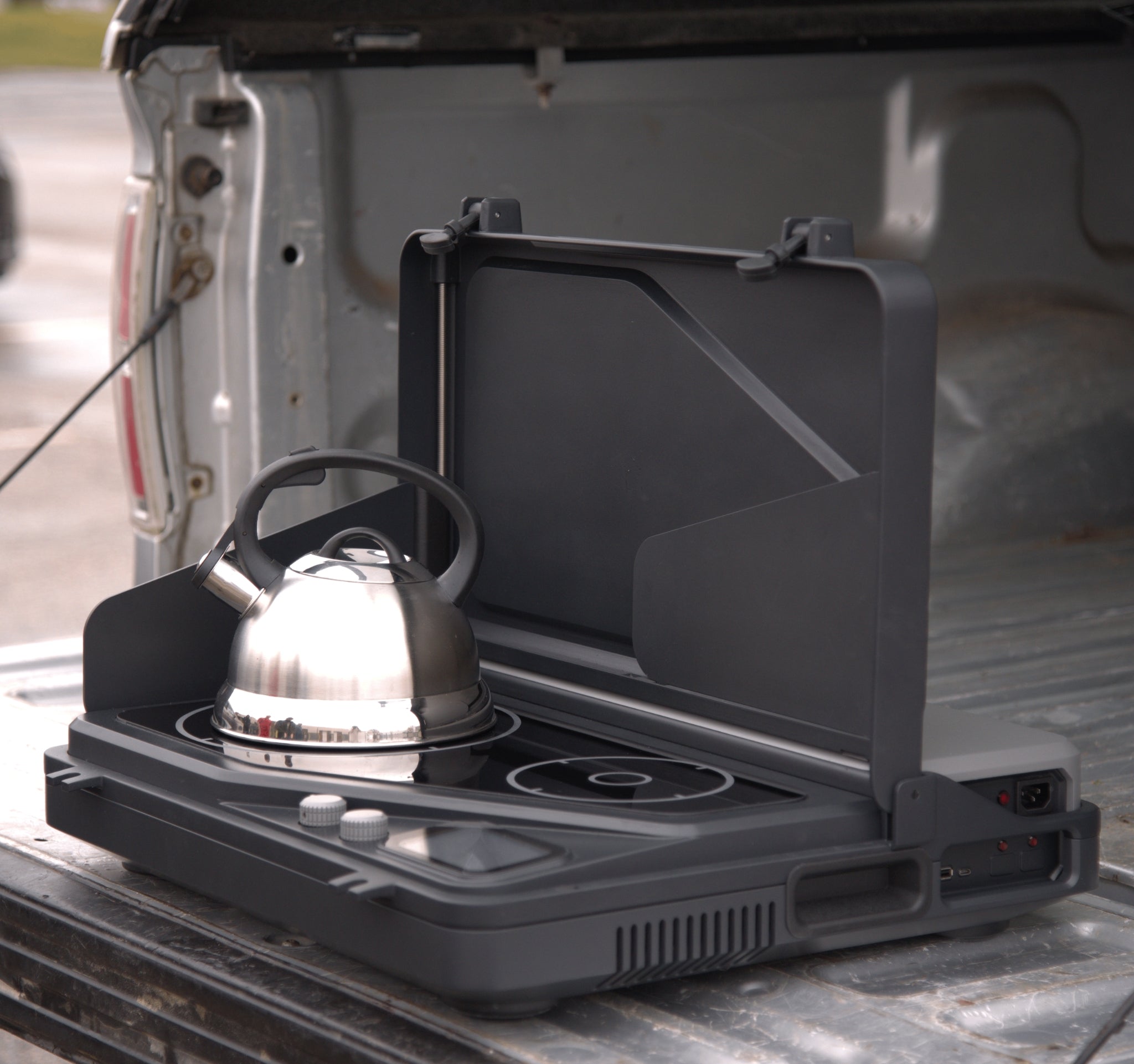 Our battery powered, portable induction cooking stove and generator, on a tailgate, for tailgating, camping, overlanding, and off-grid survival. a removable battery and dual burners for campers, outdoor workers, victims of natural disasters — and anyone, anywhere, cooking or surviving off-grid. Pictured here with the lid open with a kettle and also a coffee maker on with the dual burner system is shown in the portable Overlander model. Wind guards are currently pictured also.